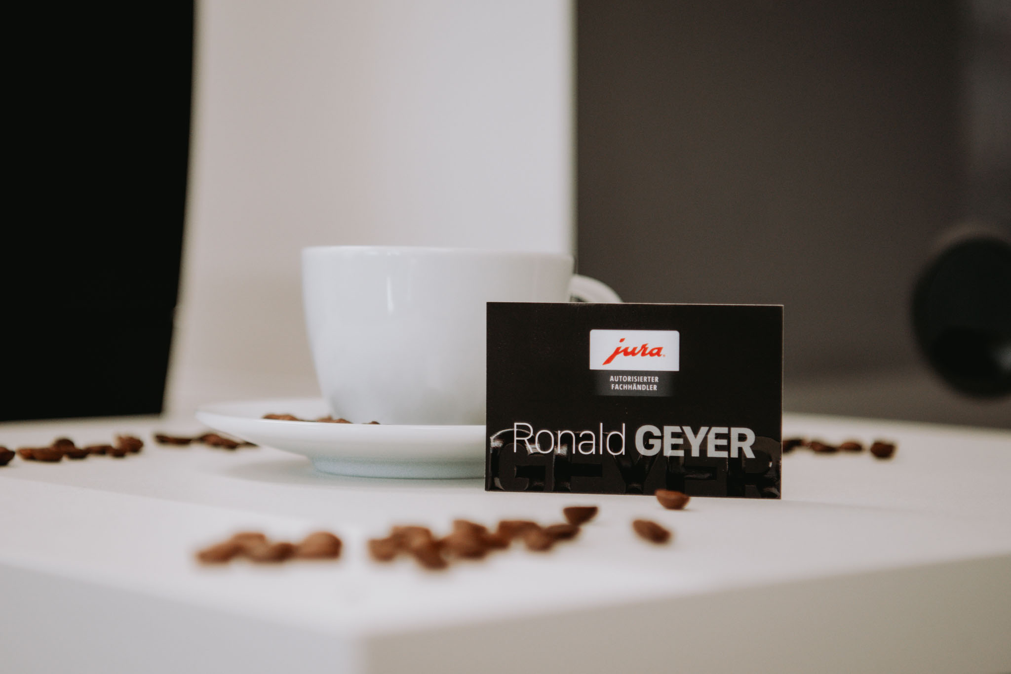 Visitenkarte von Ronald Geyer angelehnt an eine weiße Kaffeetasse und darum liegen Kaffeebohnen